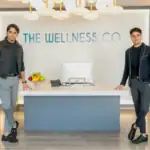 Two men standing at the reception desk of The Wellness Company, a health-focused business, with a modern interior and branding visible in the background.