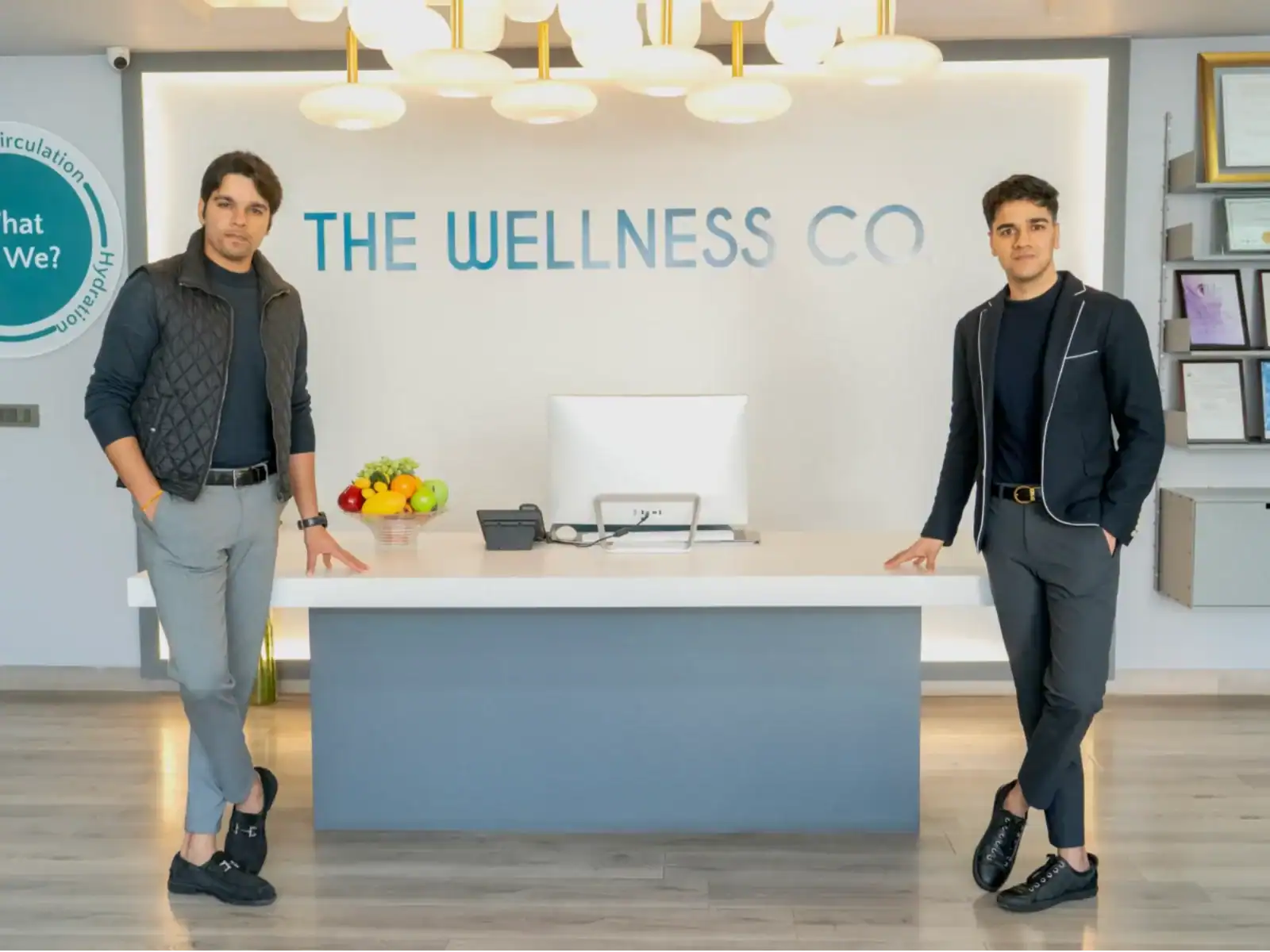 Two men standing at the reception desk of The Wellness Company, a health-focused business, with a modern interior and branding visible in the background.