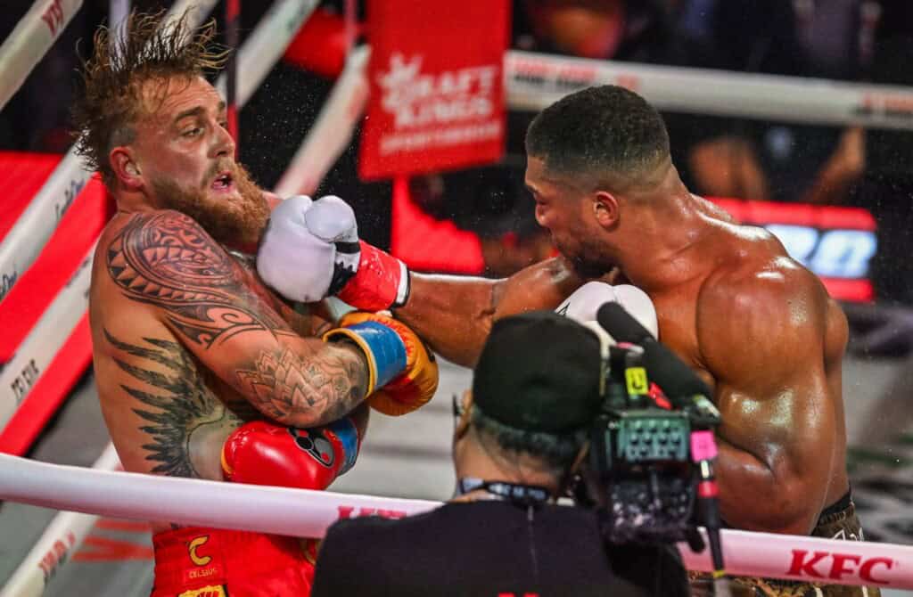 Intense boxing match between Jake Paul and Anthony Joshua with fighters exchanging punches in the ring.