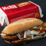Close-up of a McDonald's McRIB sandwich with sauce, onions, and pickles in front of its packaging.