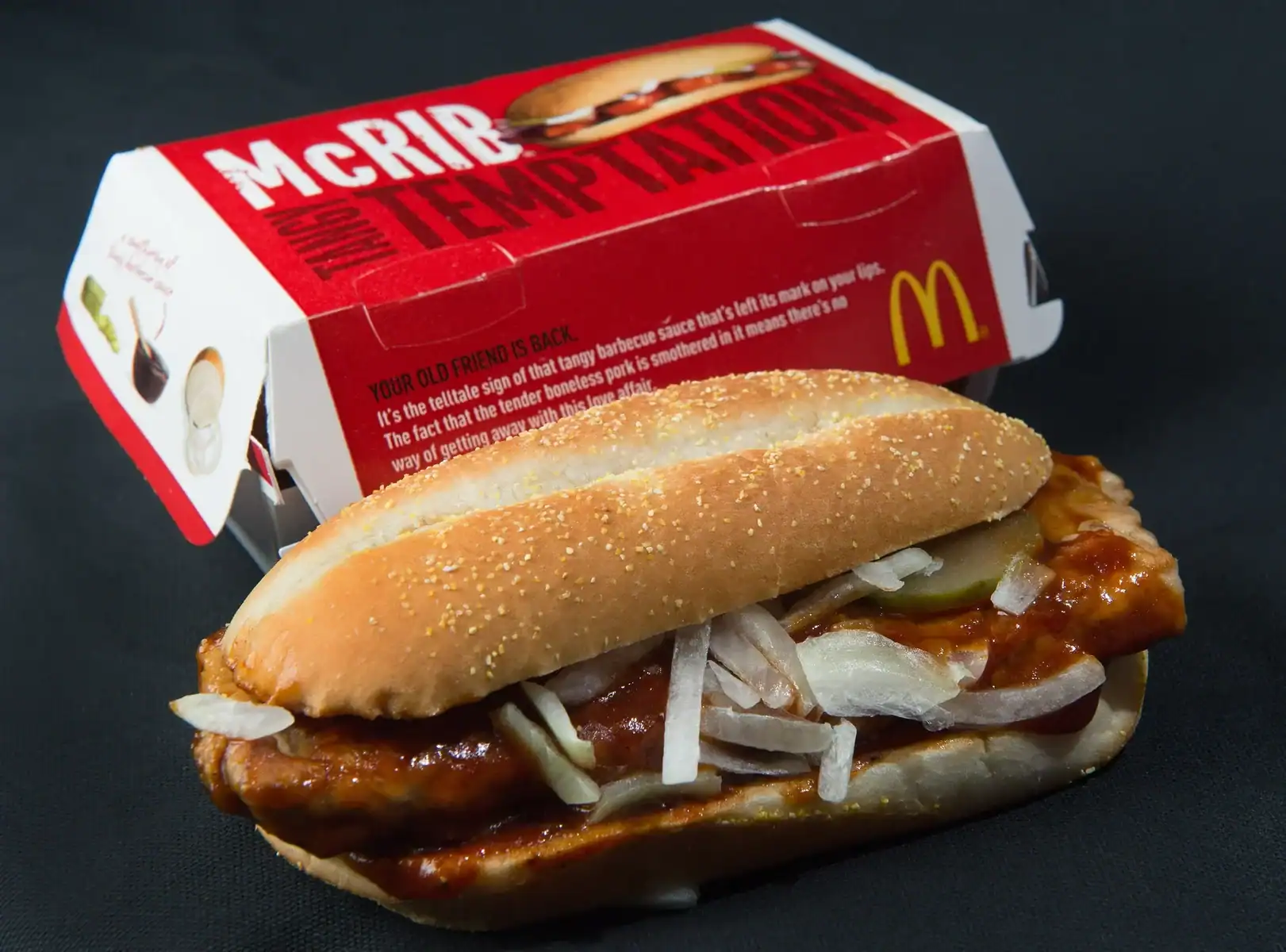 Close-up of a McDonald's McRIB sandwich with sauce, onions, and pickles in front of its packaging.