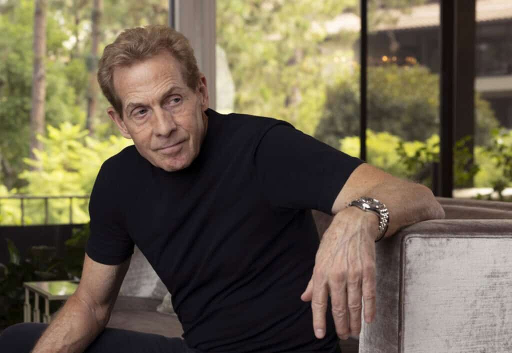 Image of Skip Bayless, sports commentator, sitting on a sofa in a modern living room, discussing legal issues related to his lawsuit.