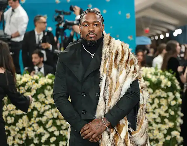 Stefon Diggs at a public event, dressed in a black suit with a fur stole, surrounded by media and fans, highlighting his legal situation regarding paternity allegations.